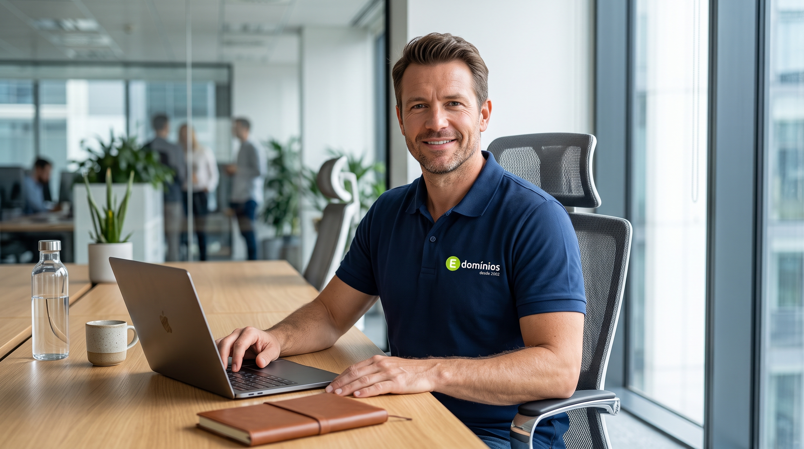 Homem sentado em uma mesa moderna de trabalho, sorrindo e usando notebook, vestindo camisa polo com o logotipo da empresa.